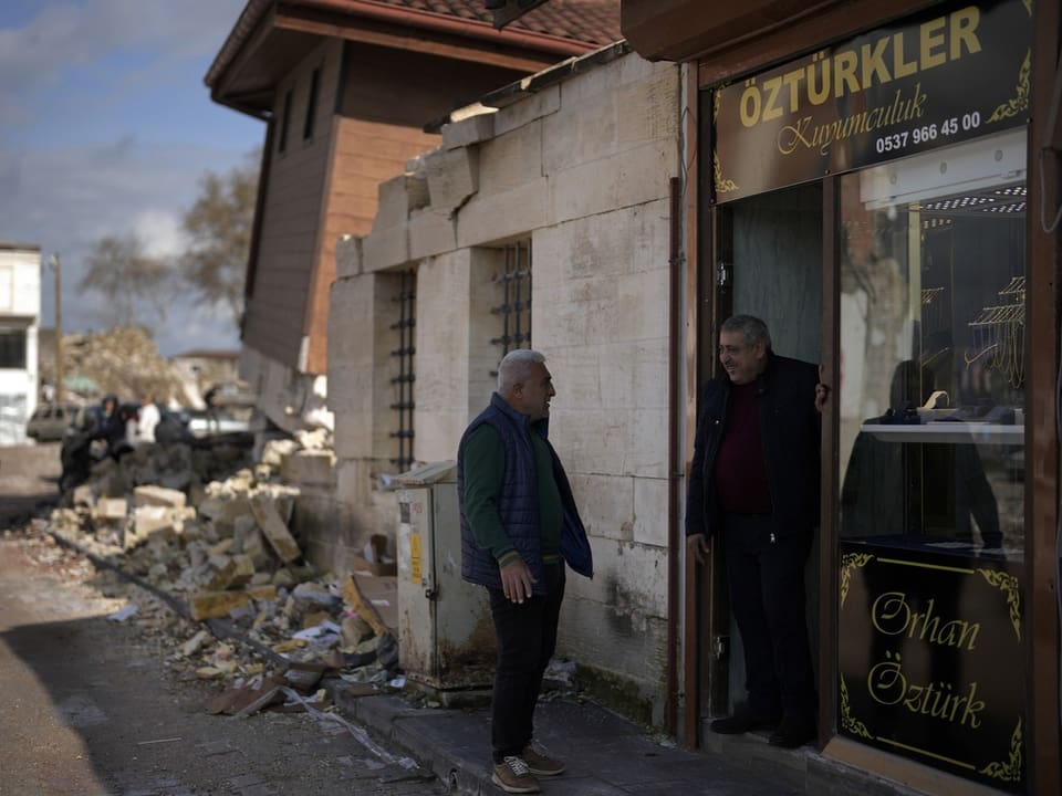 Ein Mann spricht am Eingang eines Geschäfts mit einem Freund. Dahinter sieht man Schäden des Erdbebens vor einem Jahr.