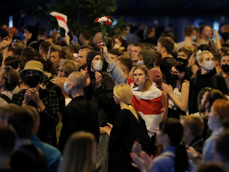 Eine Frau streckt inmitten von Demonstranten eine Hand voller Blumen in die Höhe, über die Schultern hat sie die Landesflagge von Weissrussland gehängt.
