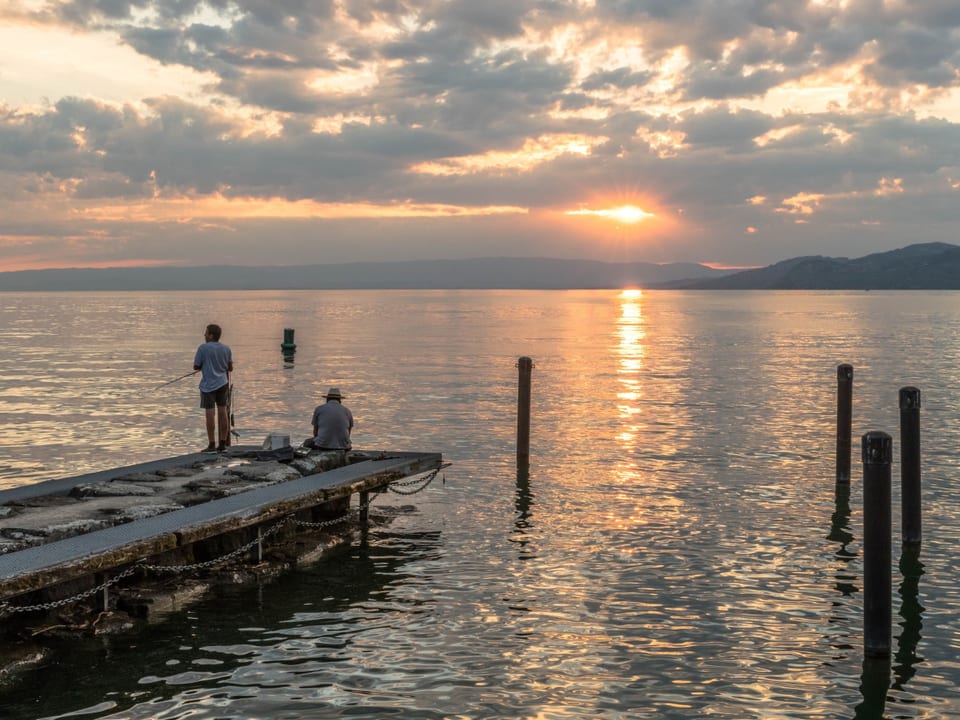 See bei Sonnenuntergang und bewölktem Himmel. am Ufer stehen zwei Fischer auf einem Steg mit Ruten in der Hand. 