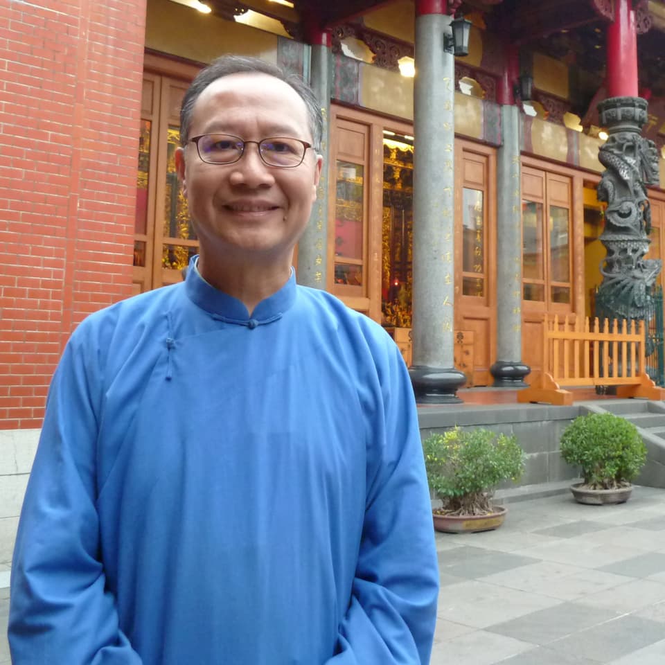 Meister Li trägt ein blaues Hemd, lächelt in die Kamer. Im Hintergrund der Eingang zum Tempel.