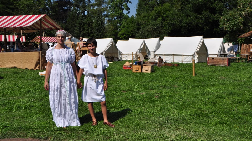Frau und junges Mädchen in Römerkleidung, weisse Tuniken und Ledersandalen. Im Hintergrund die weissen Zelte des Römerfests.