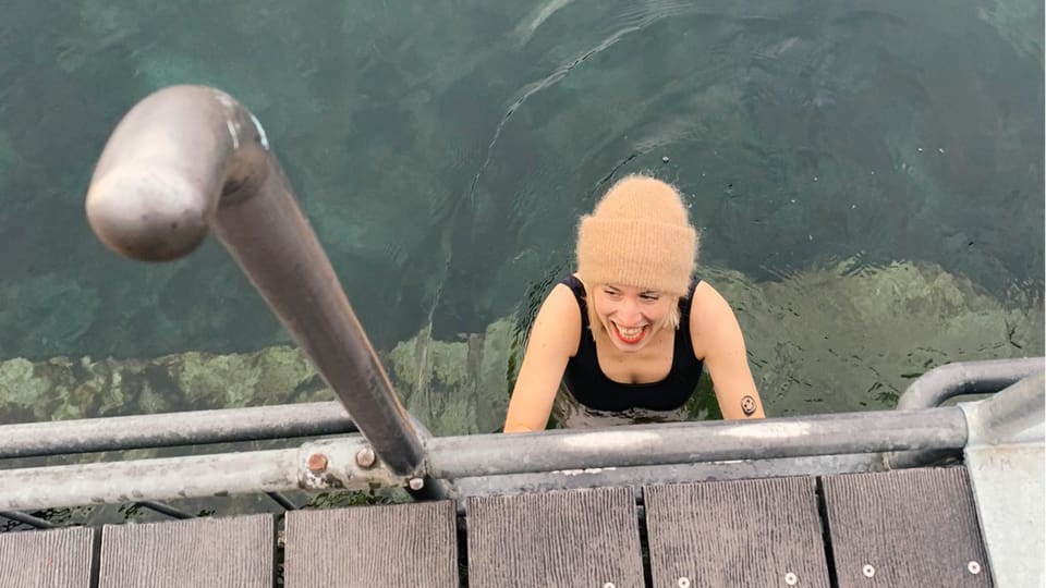 Eine blonde Frau mit Wollmütze steigt ins Wasser und lacht.