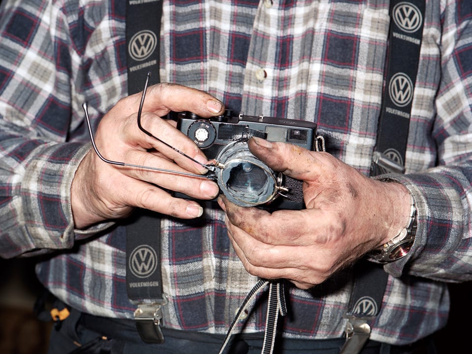 Ein älterer Mann hält eine alte analoge Kamera in der Hand. Vor der Linse hält er ein Brillenglas.