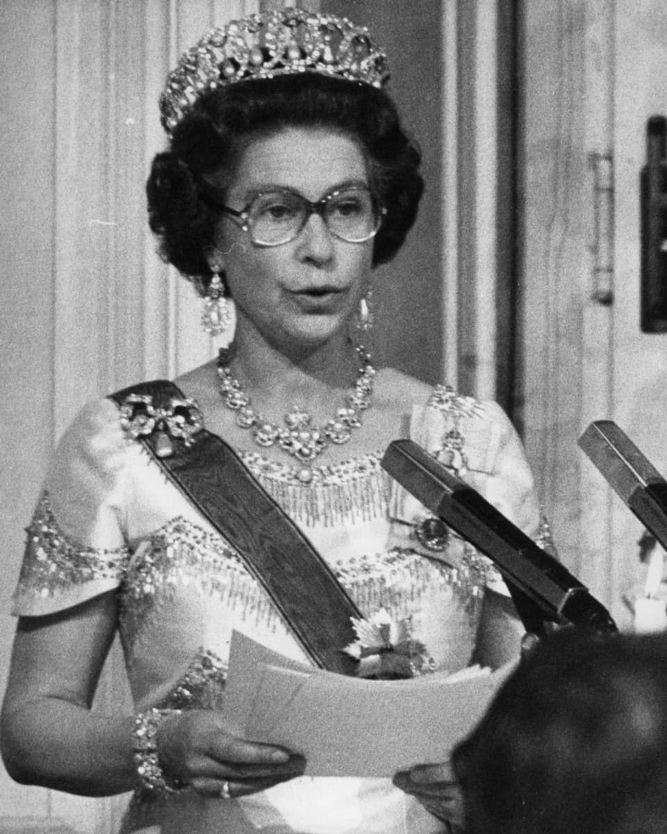 schwarzweiss Foto einer Frau mit Krone, Brille und Papieren in der Hand. Spricht in Mikrofon.