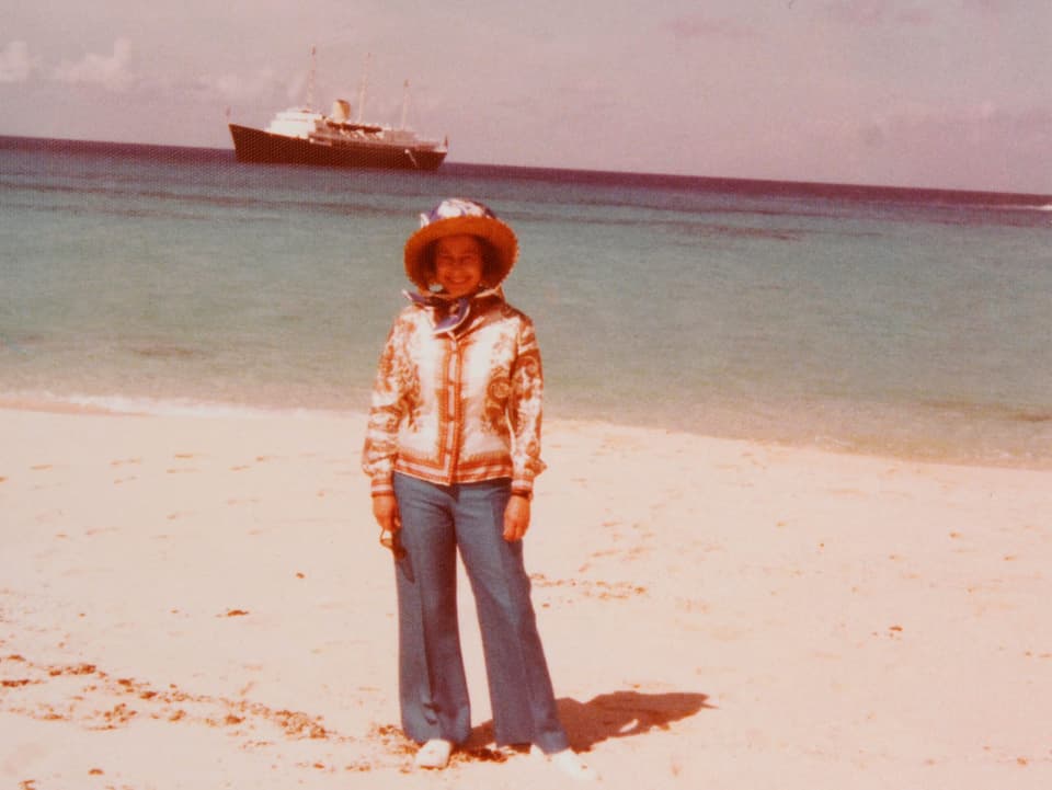 Queen Elizabethsteht mit Hosen, Blazer und Hut an einem unbekannten Strand. Hinter ihr sieht man das Meer und ein Schiff.