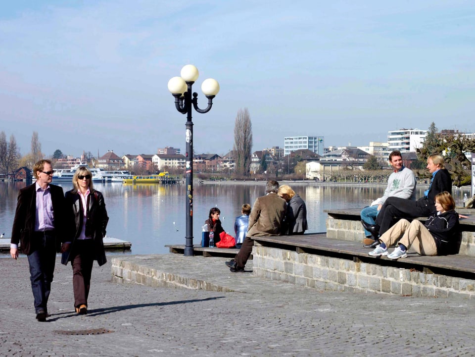 Ufer bei Sonnenschein. Es sind Menschen am spazieren und sitzen. Sie tragen Jacken und Pullover.