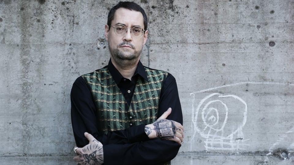 ein Mann mit vielen Tattoos an den Händen steht vor einer grauen Wand