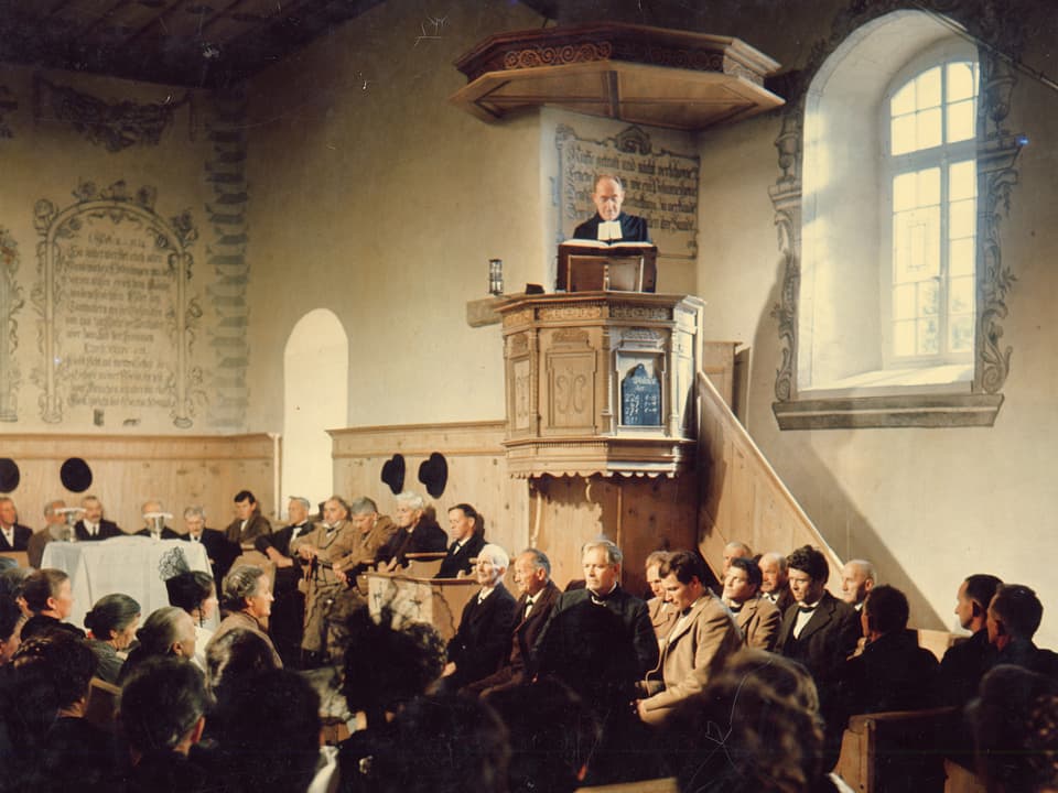 Szene in einer Kapelle. Der Pfarrrer steht in der Kanzel und predigt zum versammelten Volk.