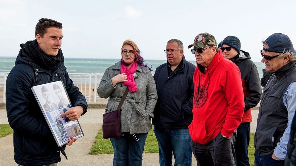 Reisegruppe steht um einen Reiseführer in der Normandie. Er erklärt etwas ab einem Plakat.