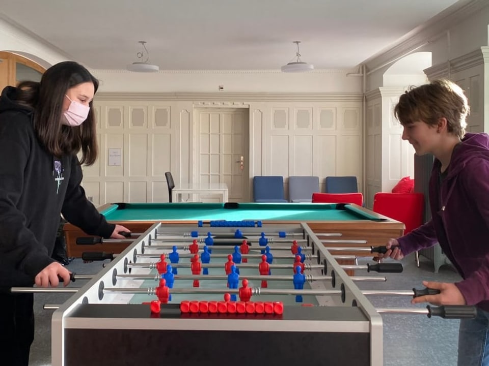 Rechts ein schwarz gekleidetes Mädchen, rechts die Kurzhaarige, spielen gemeinsam Tisch-Fussball.