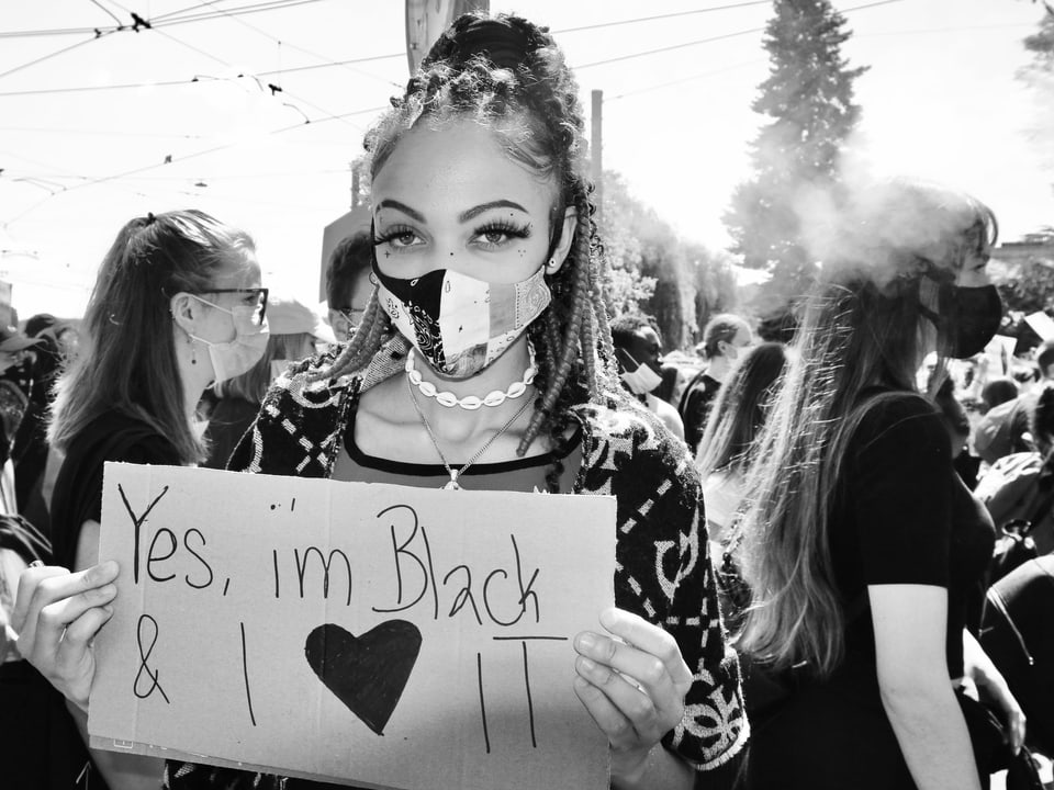 Eine junge Frau mit Dreads und Mundschutz hält ein Schild: "Yes i'm black & I love it"