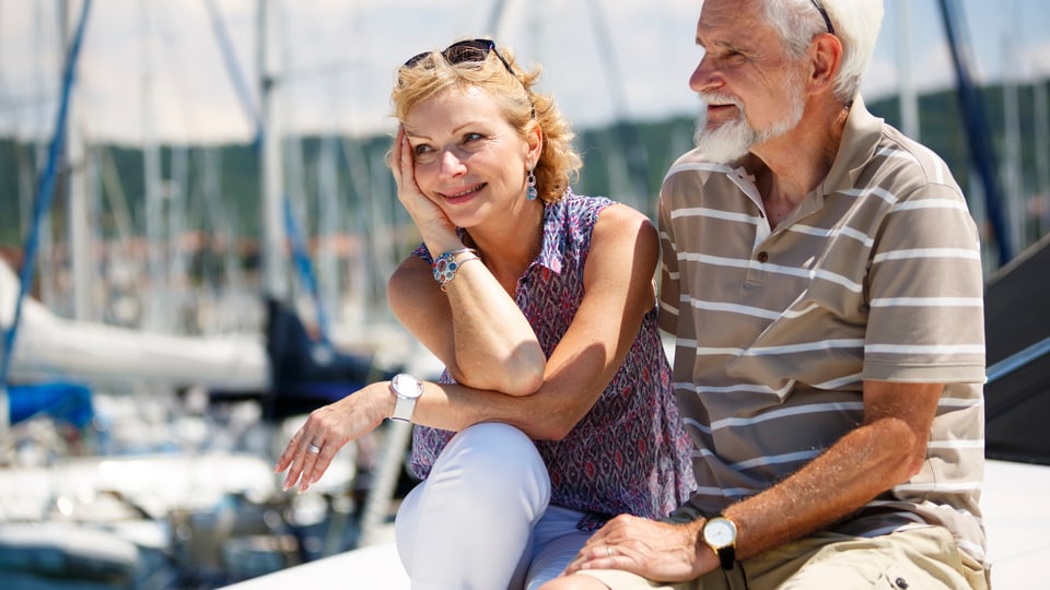 Ein älteres Ehepaar sitzt an einem Hafen und lächelt.