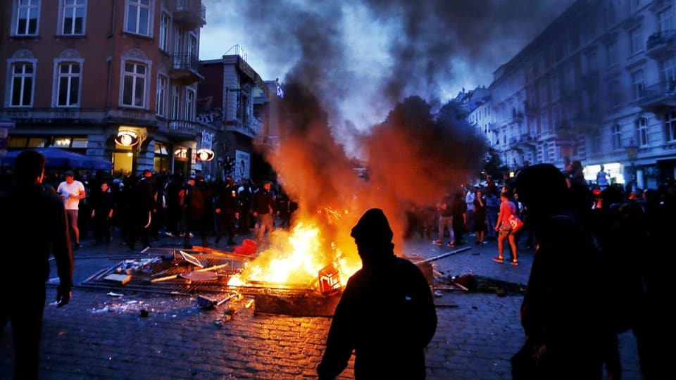 Auf einem Platz brennt ein Feuer und verschiedene Gegenstände werden verbrannt, darum stehen Menschen.