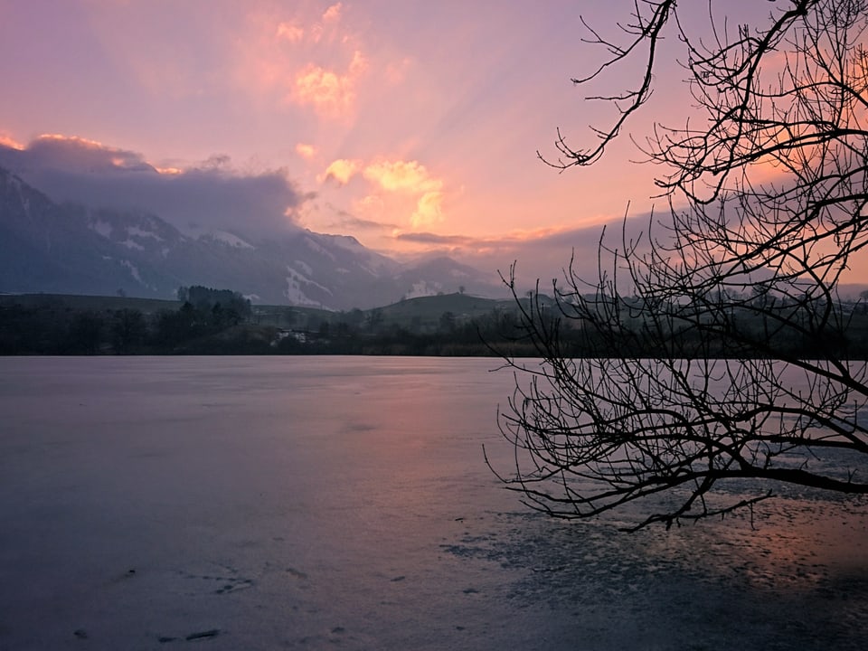 Abendstimmung in rosa und violett. Im Vordergrund ein zugefrorener See, im Hintergrund im Dunst Berge. Die Sonne versteckt sich hinter Wolken und zaubert rosa Strahlen an den Himmel.