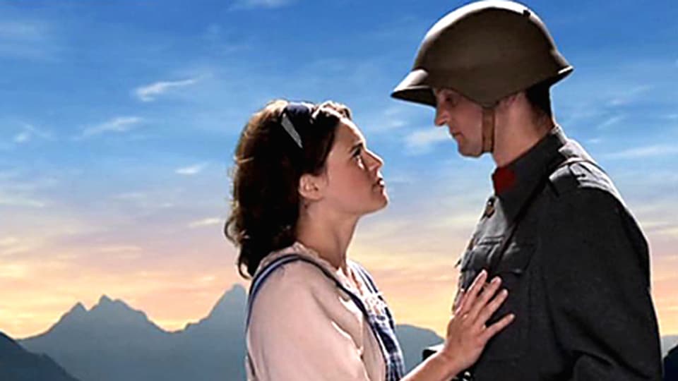Ein Soldat und seine Frau stehen sich gegenüber. Ihre Hand liegt auf seiner Brust. Im Hintergrund sieht man einen See und Berge bei Sonnenuntergang.