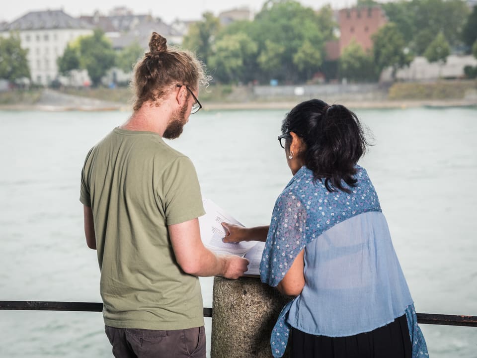 Ein Mann und eine Frau unterhalten sich beim Ufer des Rheins. Sie haben Papiere vor sich.