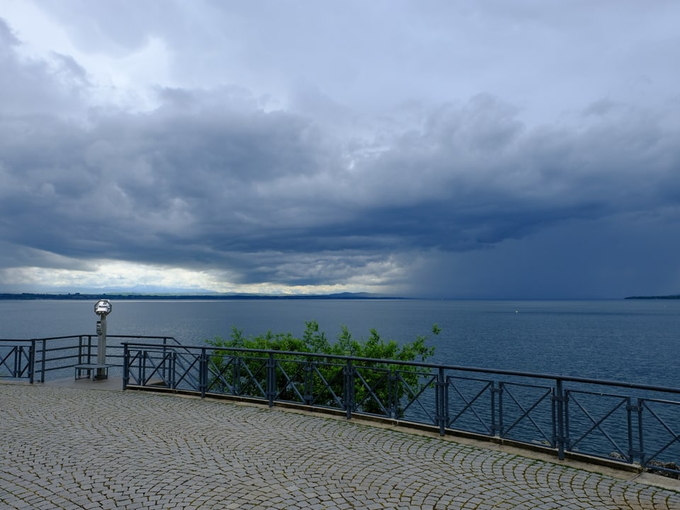 Über dem Neuenburgersee ergiesst sich aus dichten Wolken Regen, im Vordergrund eine Ausichtsplattform am Seeufer.