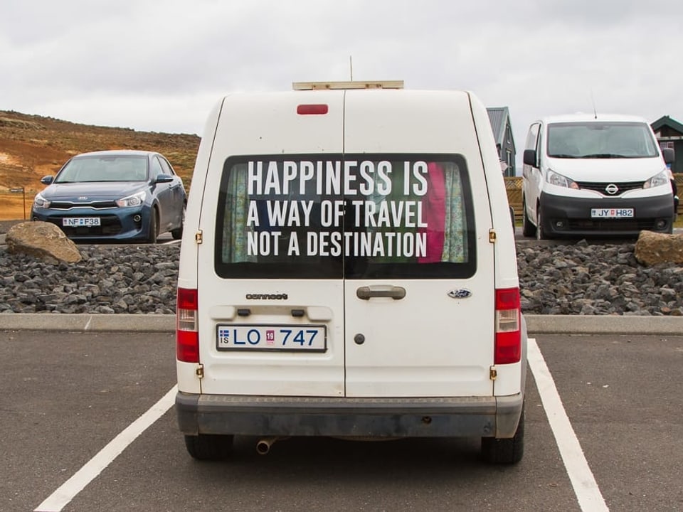 Ein Camper steht auf einem Parkplatz. Auf der Heckscheibe steht: «Happiness is a way of travel not a destination»