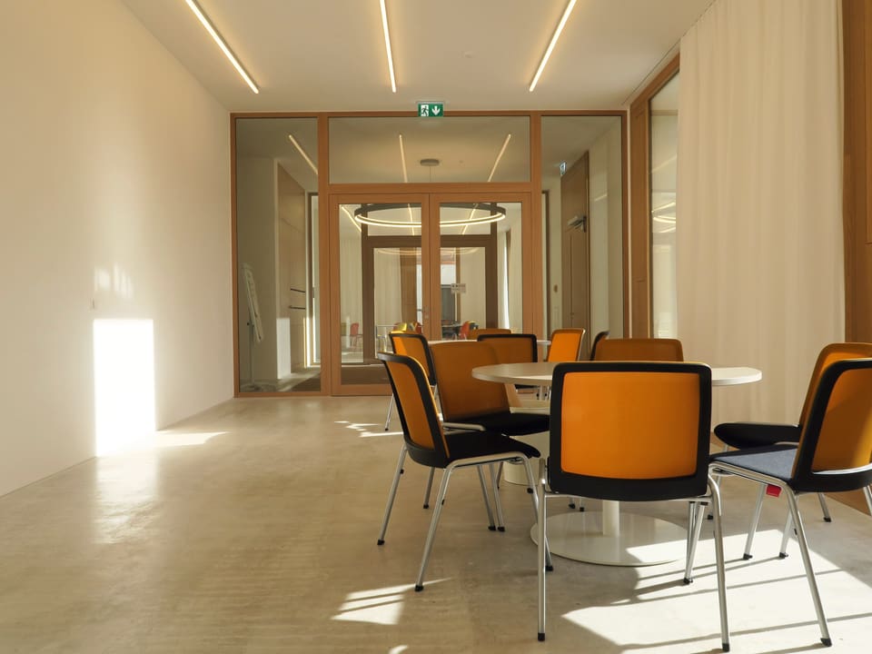 In einem hellen Raum mit viel Tageslicht stehen um einen runden weissen Tisch einige braune Stühle.