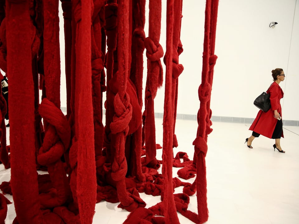 Rote Wollfäden hängen von der Decke, Frau in rotem Mantel geht daran vorbei.