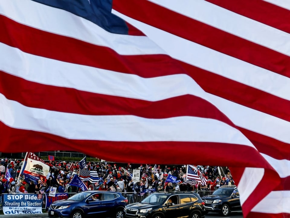 Die amerikanische Flagge ist zu sehen, darunter/dahinter Tausende Menschen, die gegen Trumps Niederlage protestieren.