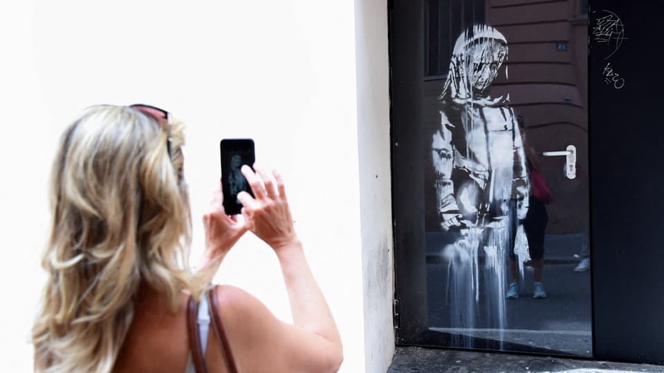 Eine Person fotografiert auf ihrem Handy eine schwarze Tür, auf die in Weiss eine traurige Madonna gesprayt ist.