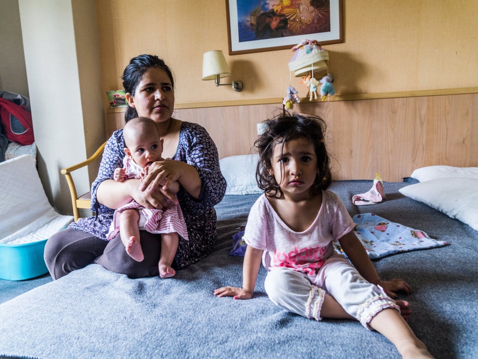 Eine Mutter sitzt mit ihren Kindern auf dem Bett eines Hotelzimmers.