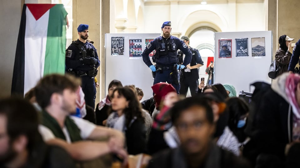 Polizeibeamte überwachen eine Menschenmenge in einer öffentlichen Halle mit Plakaten und Flaggen.