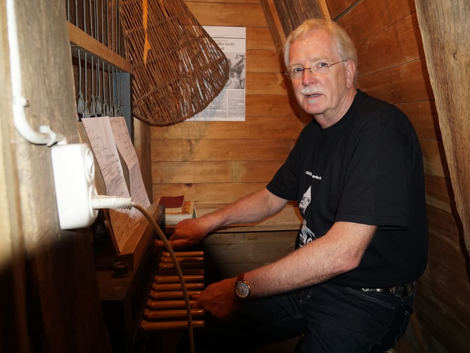 Ein Mann sitzt im T-Shirt in einer kleinen Holzkammer auf einem Stuhl - vor ihm sind elf hölzerne Hebel und Notenblätter.