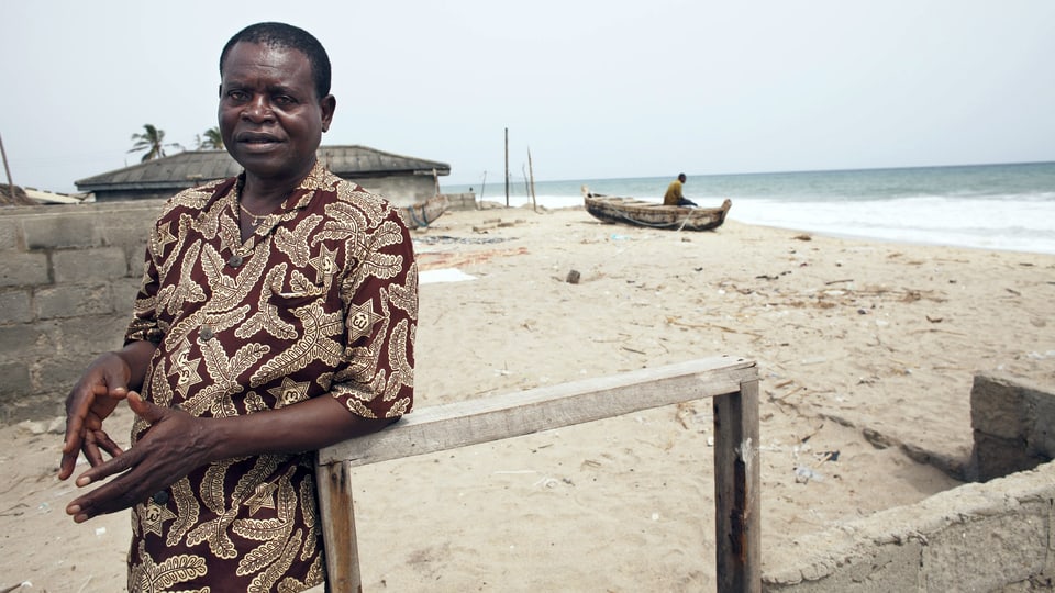 Agbakla steht vor den Ruinen seines Hauses. Im Hintergrund der Strand und das Meer.