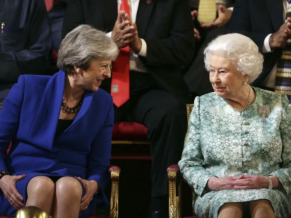 Zwei Frauen sitzen auf Stühlen. May trägt ein blaues Kostüm, die Queen einen weiss-grünes.