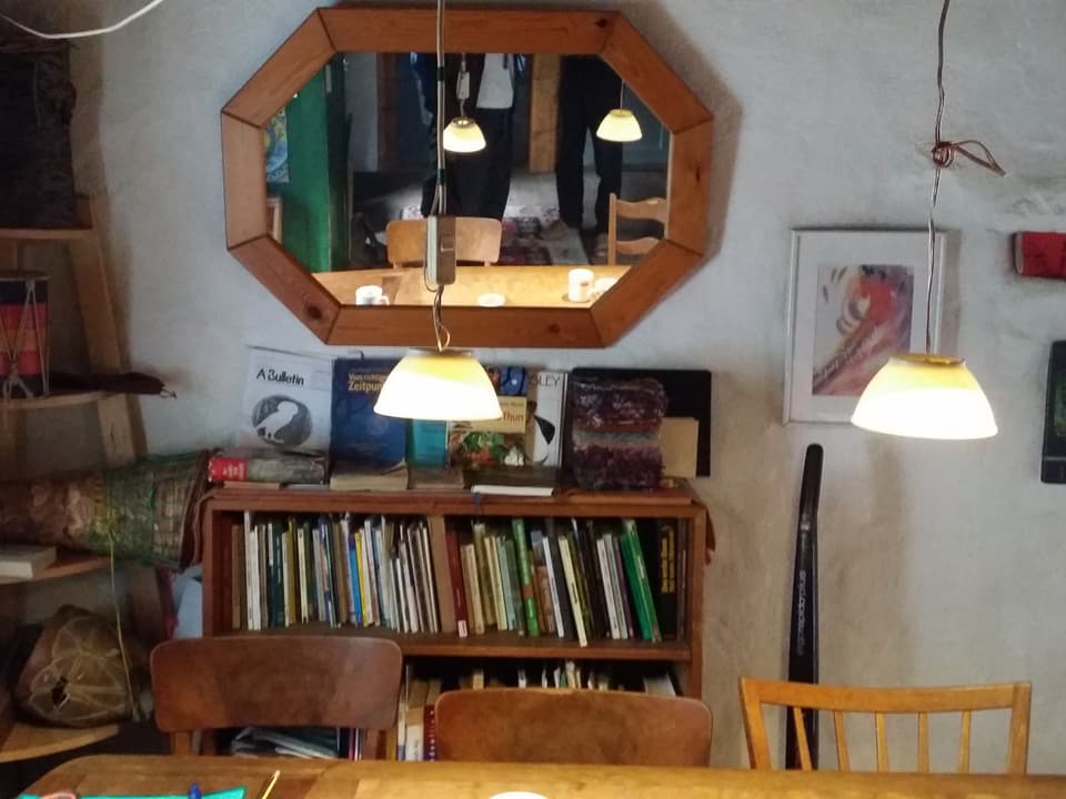 Ein Holztisch mit verschiedenen Stühlen und einem Büchergestell im Hintergrund. Zwei Lampen hangen über dem Tisch.