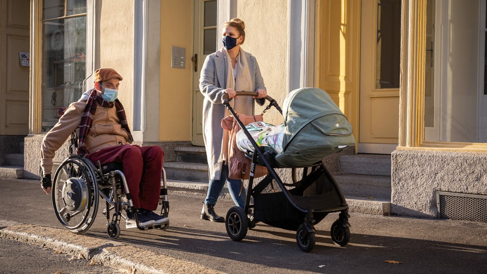 Eine Frau mit Kinderwagen spaziert auf einem Trottoir neben einem Mann im Rollstuhl.