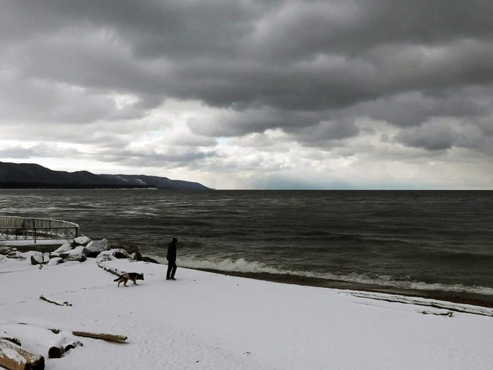 Ein Mensch steht am Ufer or dem sibirischen Baikalsee, der Himmel ist wolkenbehangen. 