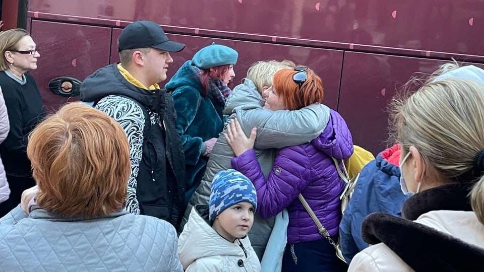 Zwei Frauen umarmen sich vor einem Bus, daneben stehen mehrere Personen.