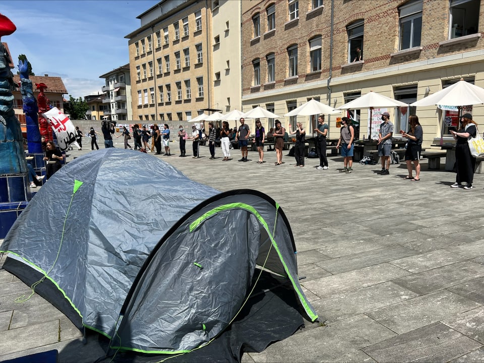 Menschen in einer Reihe mit einem Zelt im Vordergrund auf einem Platz.
