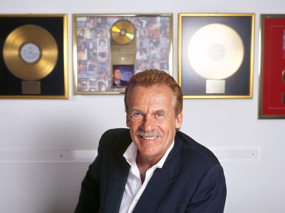 Ein Mann im weissen Hemd und dunklen Jackett sitzt vor einer Wand, an der goldene Schallplatten hängen.