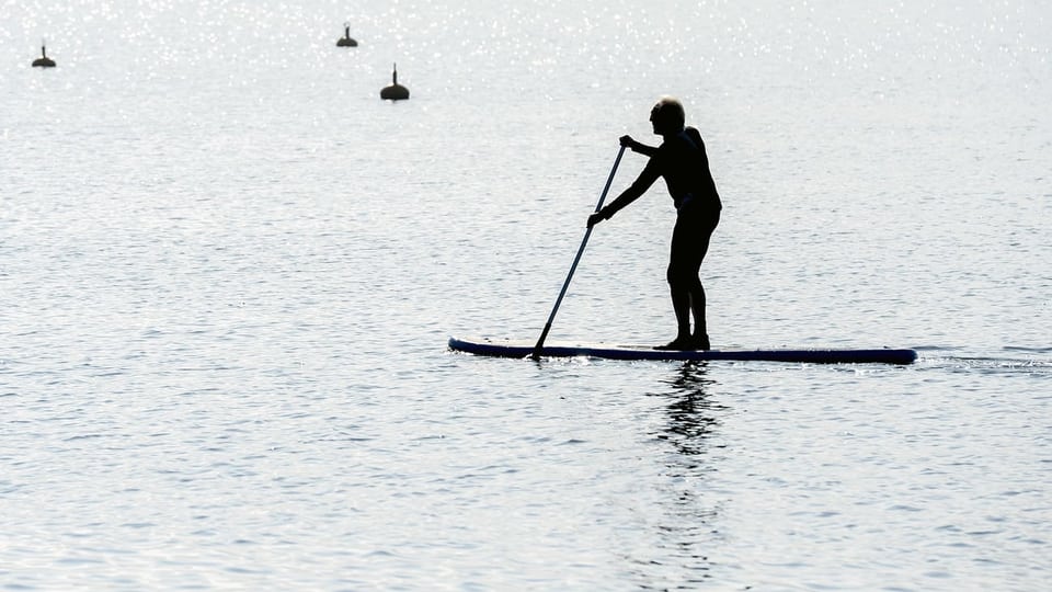 Ein Mann steht auf einem Brett, einem Stand-Up-Paddle auf dem Pfäffikersee. In der Hand hält er ein Paddel.