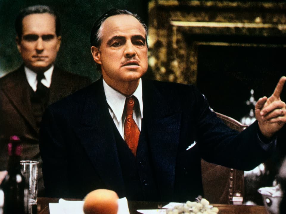 Vito Corleone sitzt an einem Esstisch und gestikuliert.