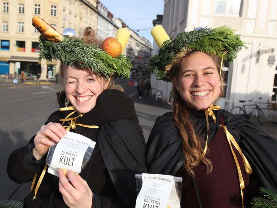 Zwei Frauen mit Adventskränzen auf dem Kopf und schwerzen Kleidern lachen in die Kamera.