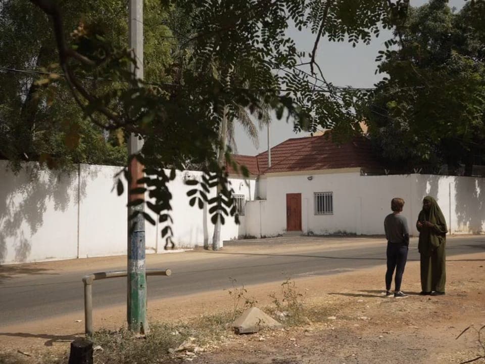 Fatoumatta und die Reporterin stehen vor einem Gebäude. Es ist hinter weissen Mauern versteckt.
