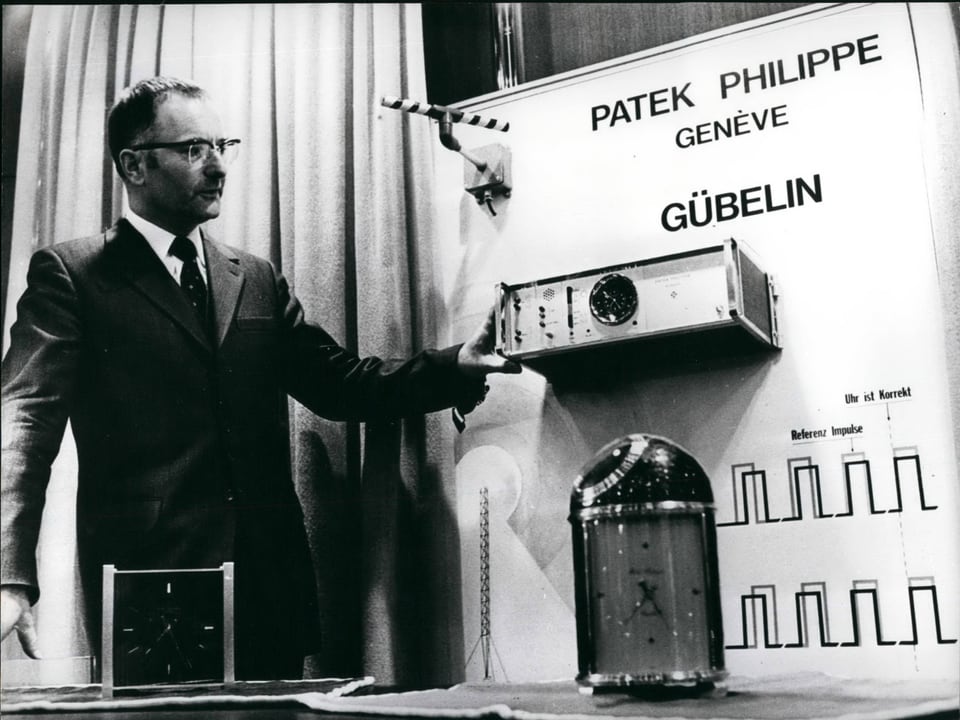 Ein Mann präsentiert eine weckergrosse Quarz-Uhr, die mit einer Atomuhr im Hintergrund via Radiowellen verbunden ist.