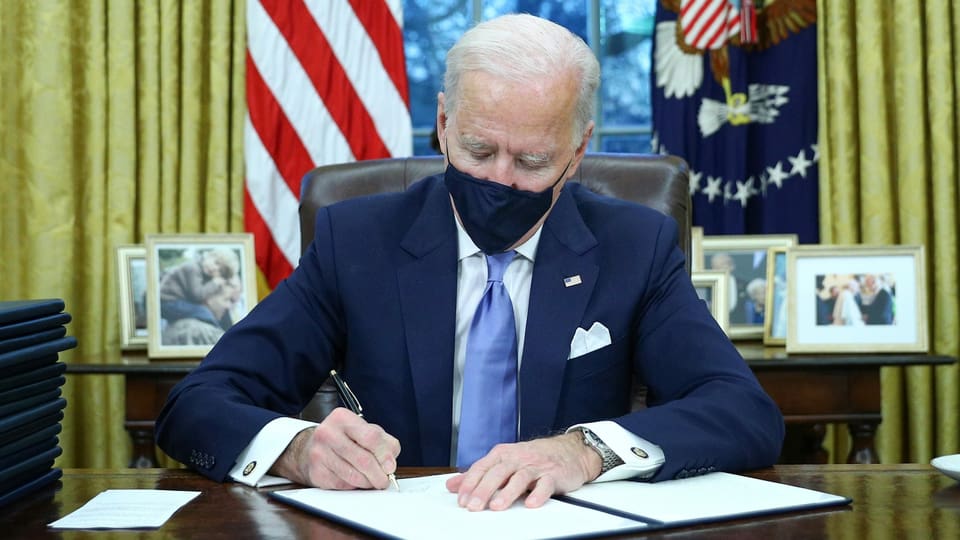 Mann mit Maske unterschreibt ein Dokument. Dahinter US-Fahnen.