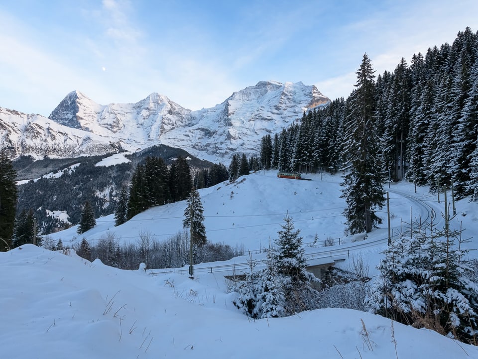 Die Bahn fährt im Berner Oberland durch die verschneite Landschaft, im Hintergund die Alpengipfel, darüber ein teils klarer und teils bewölkter Himmel.
