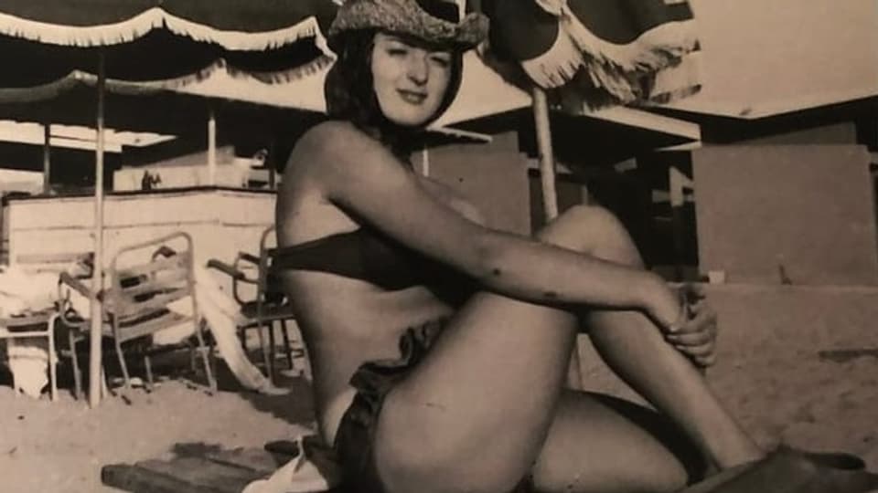 Eine junge Frau sitzt im Badeanzug mit Hut am Strand.