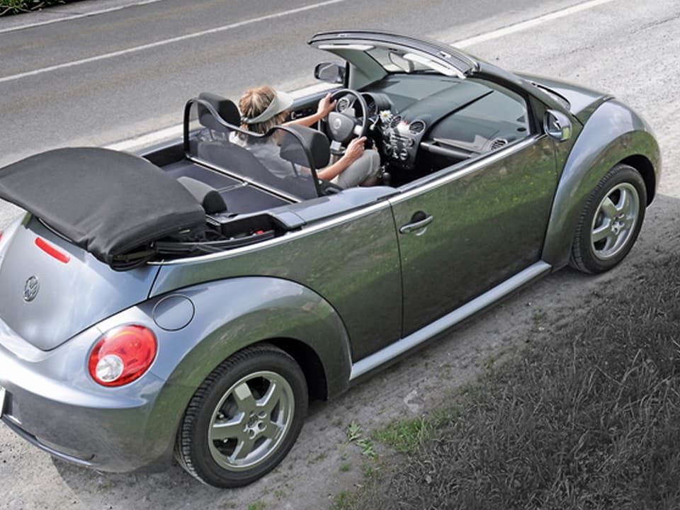 Ein hellgrauer VW-Käfer steht mitsamt Fahrerin darein, am Strassenrand.