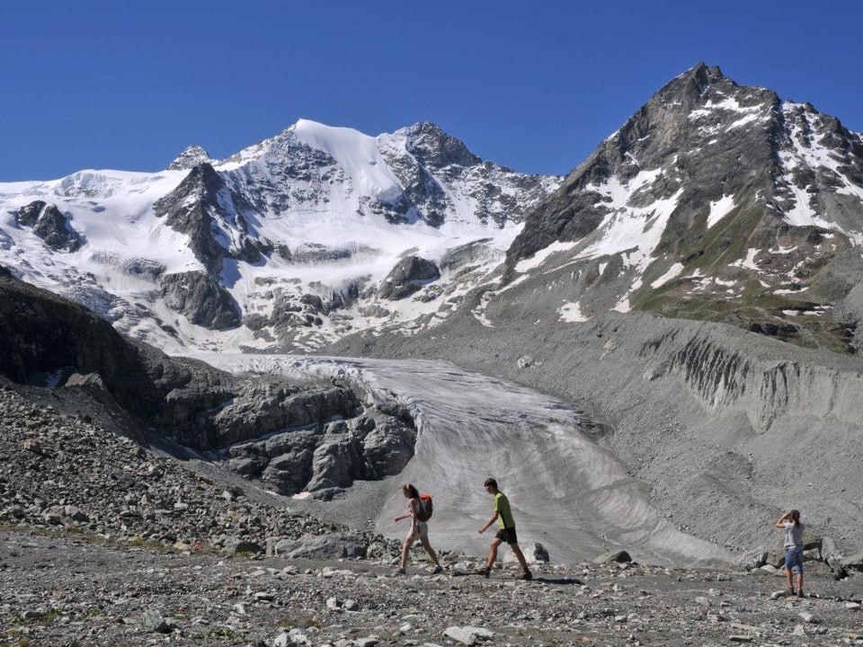 Felsiges Hochgebirge mit drei Wanderern. Auf den Gipfeln liegt immer noch Schnee, es scheint trotzdem heiss zu sein. 