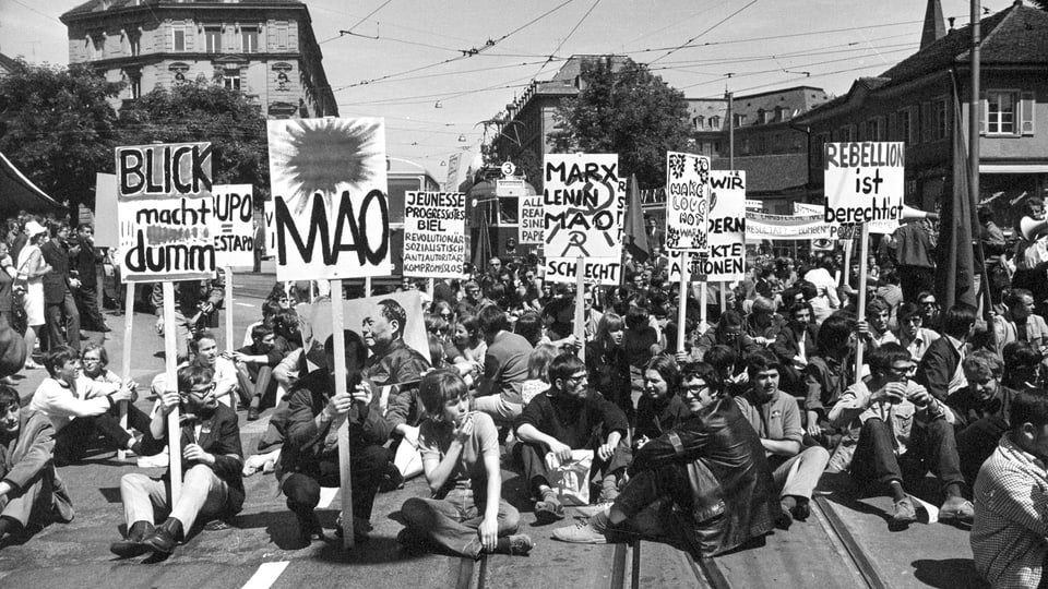 Junge Hippies sitzen auf der Strasse mit Schildern in der Hand.