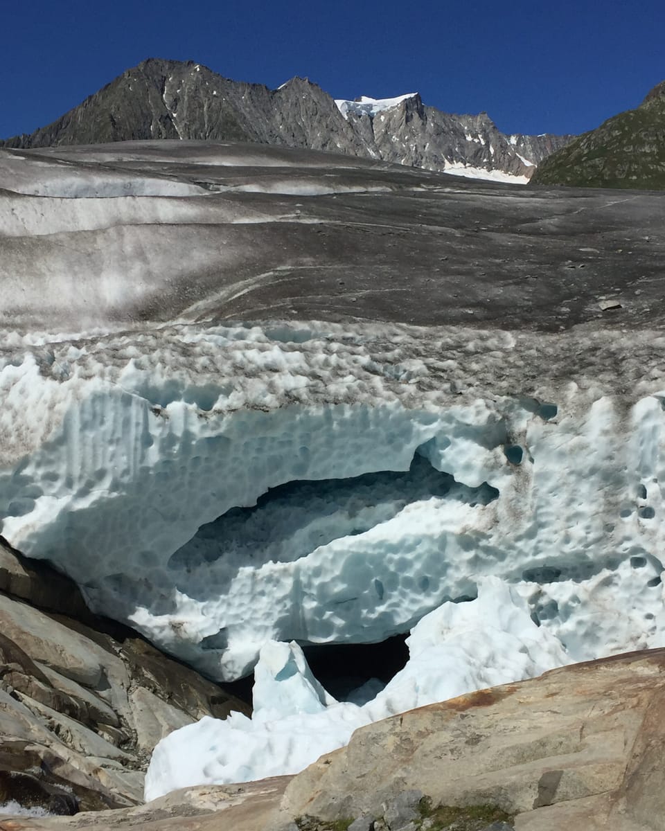 Der Himmel ist tiefblau, die umliegenden Berge und die Gletscheroberfläche grau oder mindestens gräulich. Der Betrachter steht am Eingang des Gletschers. Der Blick in den Gletscher offenbart einige Löcher ung Gruben.