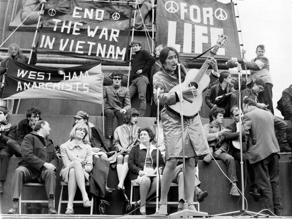 Plakate mit Kriegsprotesten, davor sitzen viele Leute und eine Frau steht mit einer Gitarre vor einem Mikrofon